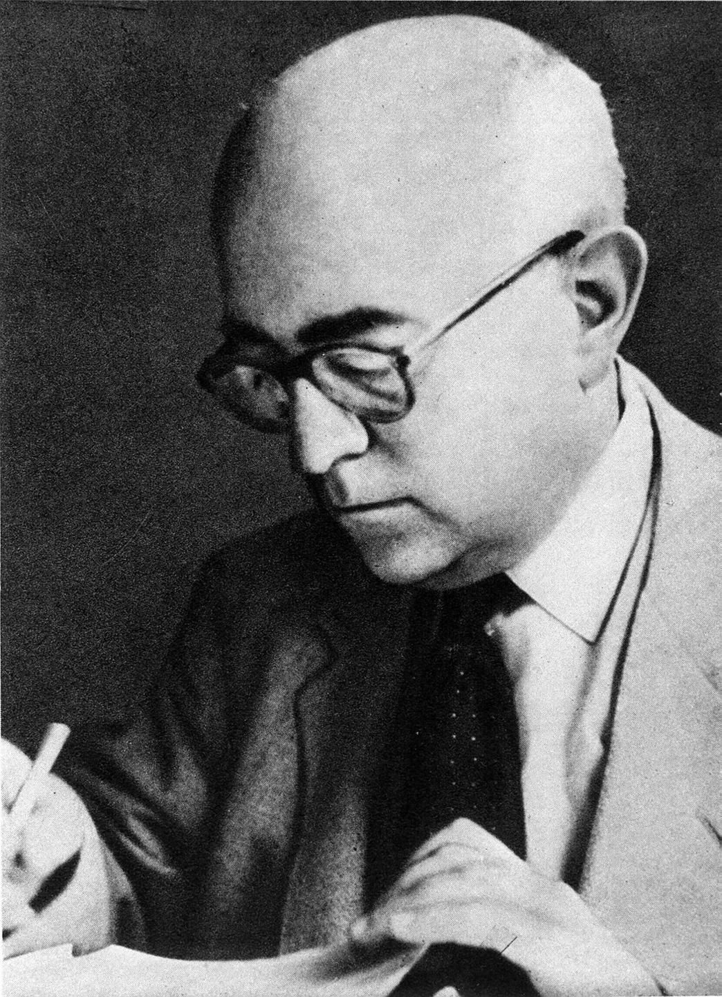 Theodor W. Adorno, im hellgrauen Sakko, mit weißem Hemd und dunkler Krawatte, schaut konzentriert auf ein Blatt Papier und schreibt.