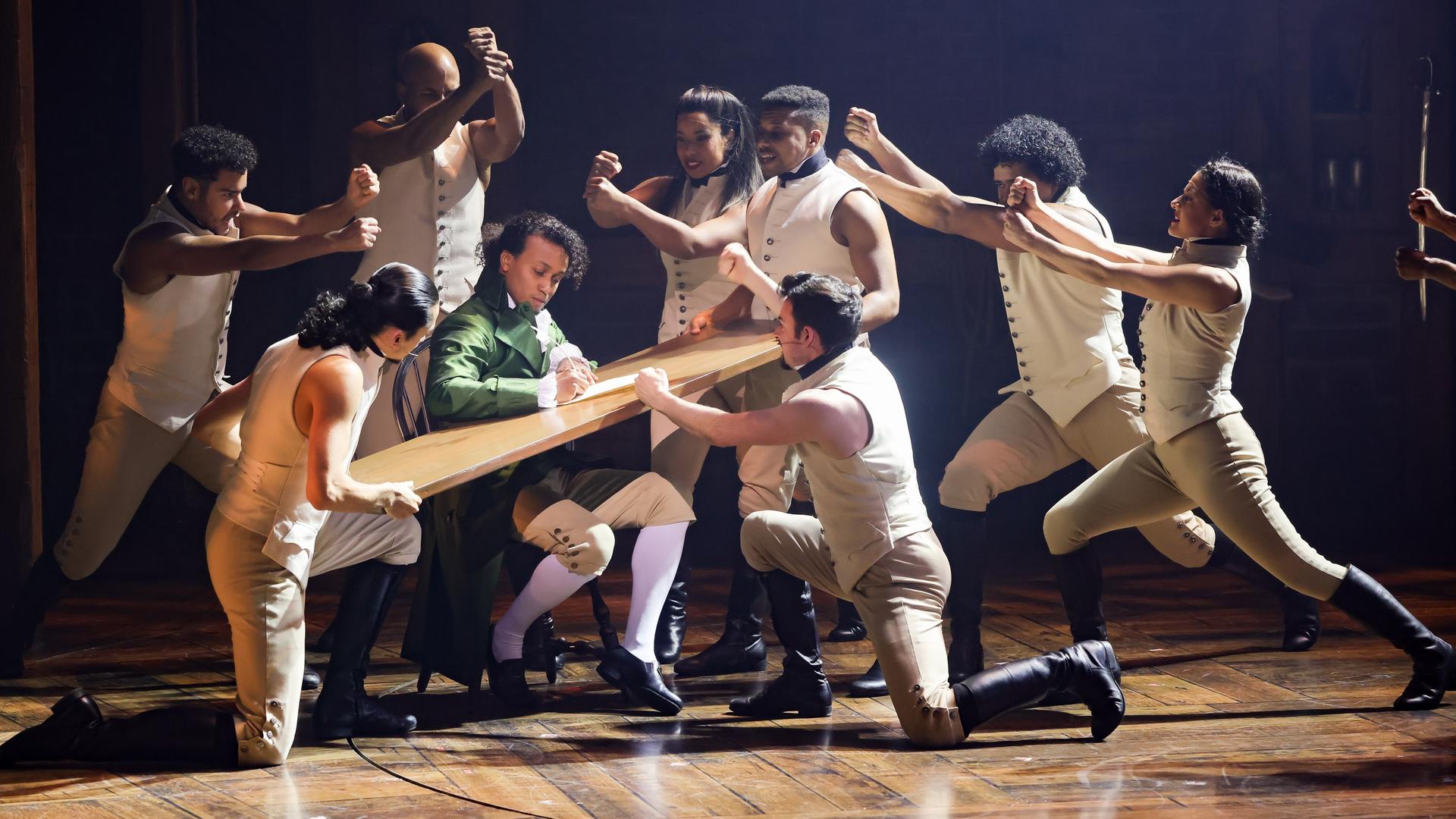 Szene aus dem Musical "Hamilton" im Operettenhaus Hamburg: Mehrere Tänzerinnen und Tänzer gruppieren sich um eine Holzplatte, die Benét Monteiro in der Hauptrolle als Schreibunterlage dient. Szene aus dem Musical "Hamilton" im Operettenhaus Hamburg: Mehrere Tänzerinnen und Tänzer gruppieren sich um eine Holzplatte, die Benét Monteiro in der Hauptrolle als Schreibunterlage dient.
