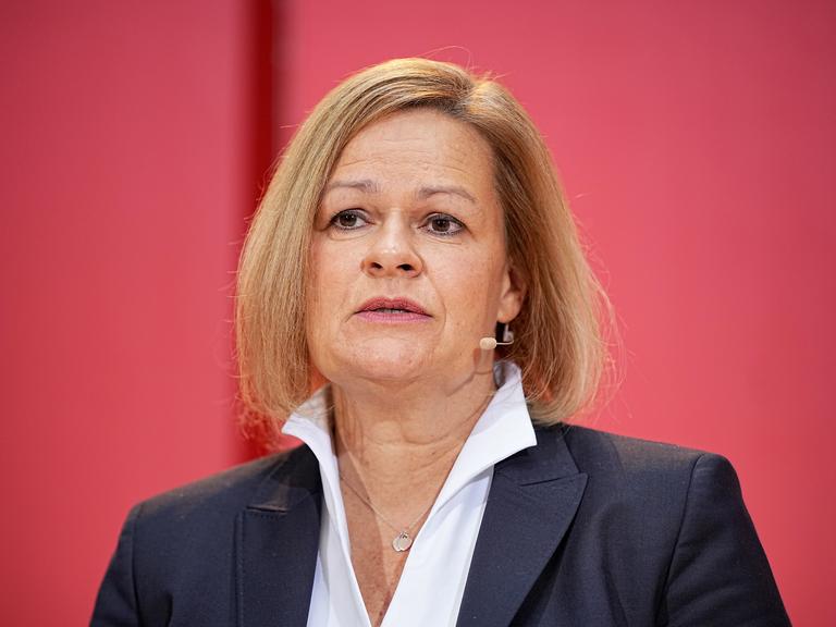 Nancy Faeser mit dunklem Jackett und weißem Hemd vor einer roten Wand.