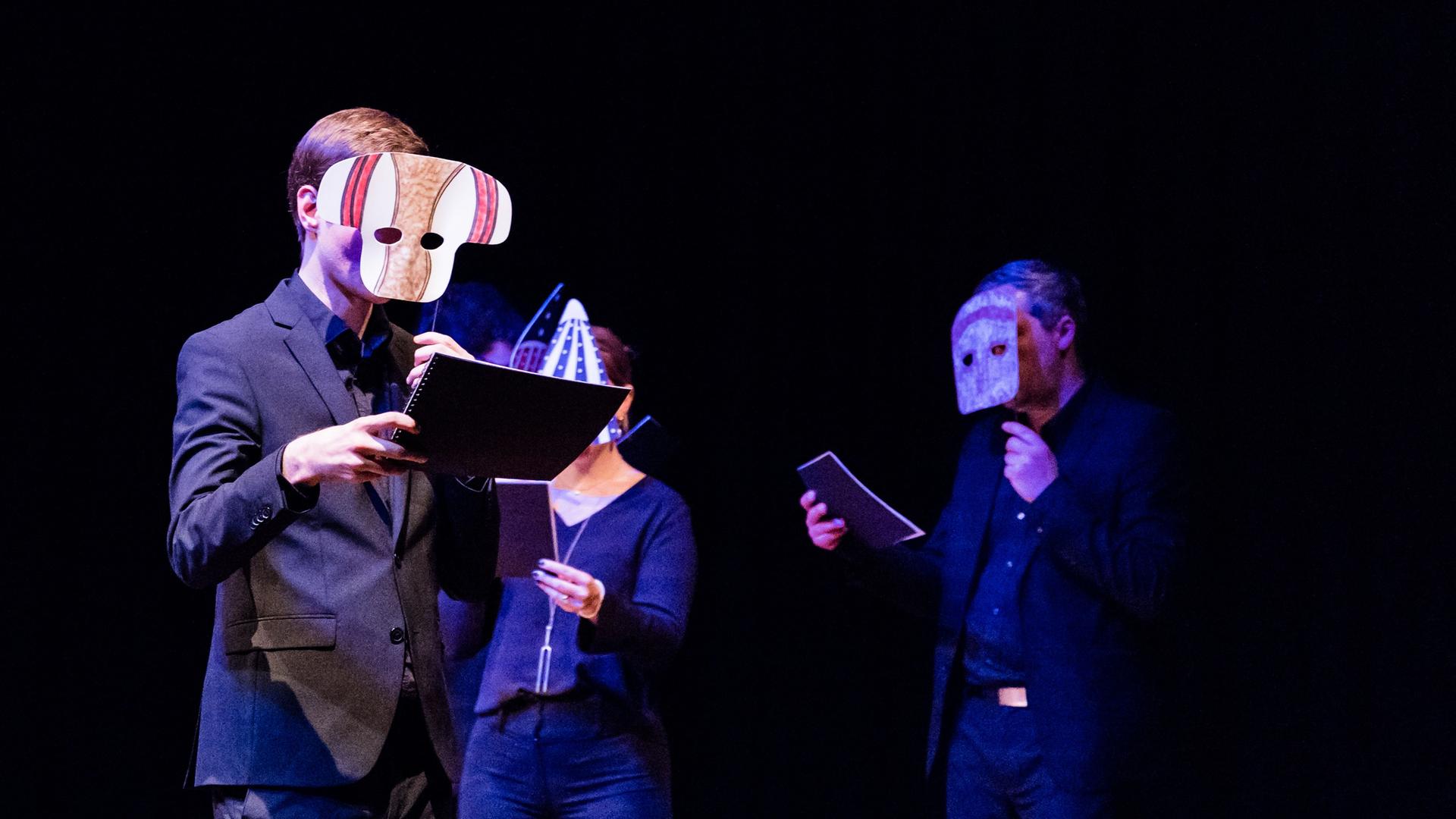 Drei Sänger und Sängerinnen gehen durch einen dunklen Raum und halten Masken vor ihre Gesichter. Drei Sänger und Sängerinnen gehen durch einen dunklen Raum und halten Masken vor ihre Gesichter.