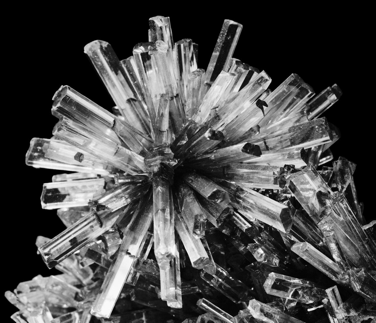 Nahaufnahme einer kugelförmigen Kristall-Formation, deren röhrenartige, durchsichtige Einzelkristalle strahlenförmig in alle Richtungen auseinander streben. (Schwarz-weiß-Fotografie)
