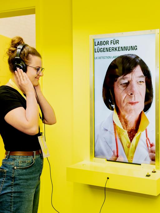 Man sieht eine Frau mit aufgesetztem Kopfhörer vor gelbem Hintergrund an einer Station mit der Aufschrift "Labor für Lügenerkennung".
