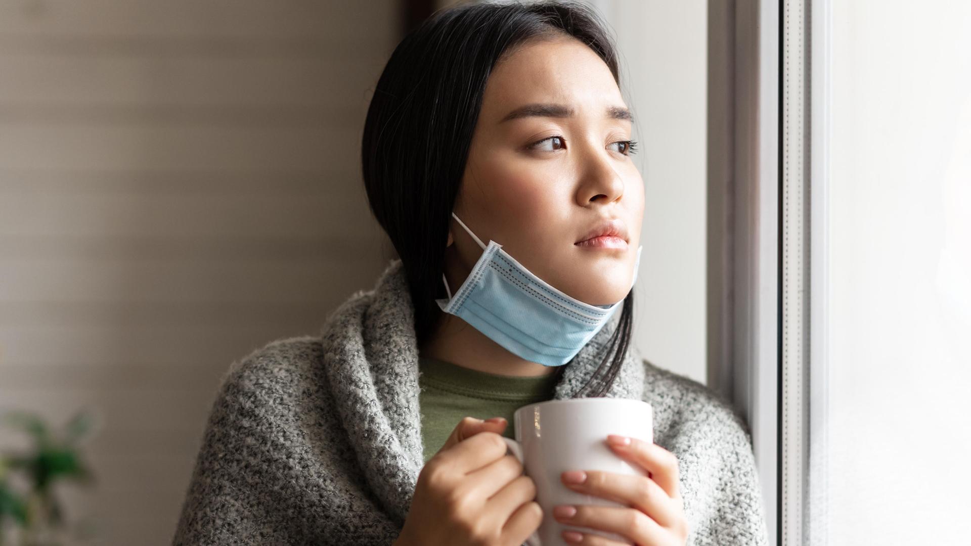 Eine junge asiatische Frau mit heruntergezogenen Mundschutz und einer Tasse in der Hand. Eine junge asiatische Frau mit heruntergezogenen Mundschutz und einer Tasse in der Hand.