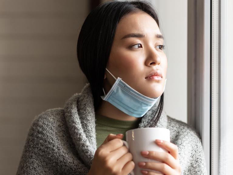 Eine junge asiatische Frau mit heruntergezogenen Mundschutz und einer Tasse in der Hand. 