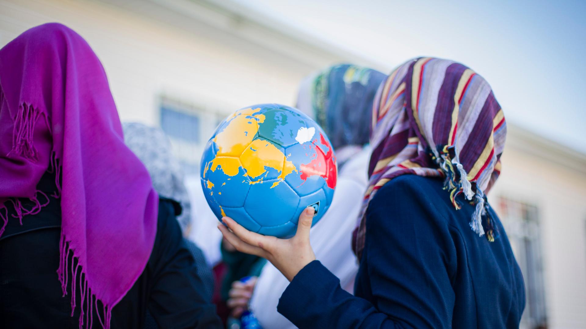 Ein syrisches Mädchen mit Kopftuch hält in einem Flüchtlingscamp einen Fussball in Form einer Weltkugel in den Händen. Ein syrisches Mädchen mit Kopftuch hält in einem Flüchtlingscamp einen Fussball in Form einer Weltkugel in den Händen.