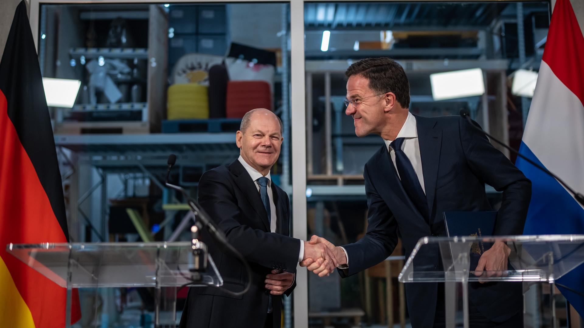 Scholz und Rutte stehen hinter ihren Rednerpulten und schütteln sich die Hände. Rutte schaut Scholz an, dieser sieht in die Kamera. Links und rechts sind die Flaggen von Deutschland und den Niederlanden zu sehen. Scholz und Rutte stehen hinter ihren Rednerpulten und schütteln sich die Hände. Rutte schaut Scholz an, dieser sieht in die Kamera. Links und rechts sind die Flaggen von Deutschland und den Niederlanden zu sehen.