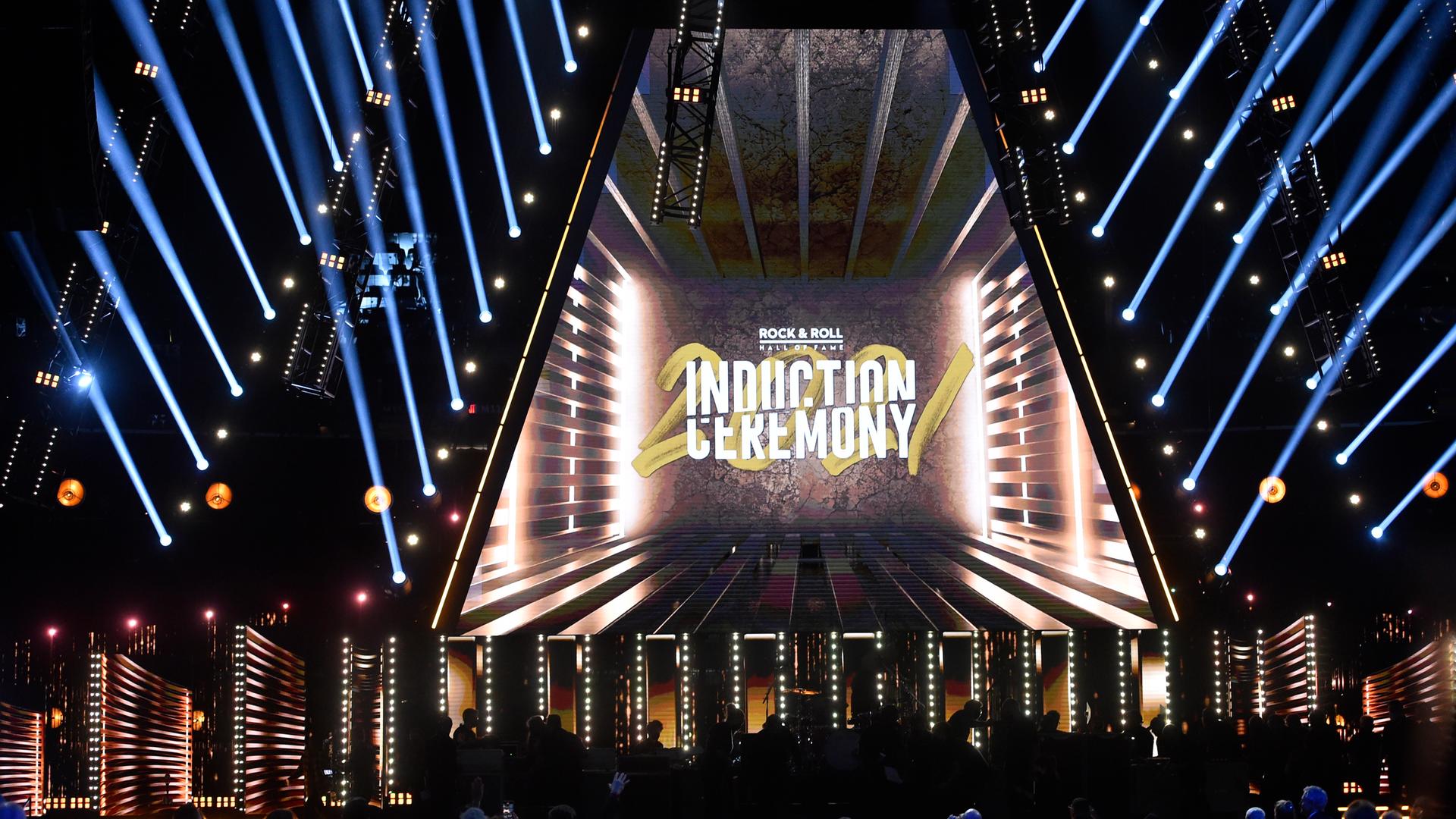 Eine leere Konzertbühne wird durch eine Lichtprojektion erleuchtet, an den Seiten werfen jeweils acht Punktstrahler blaue Lichtkegel in den Saal. In der Mitte steht "Rock & Roll Hall of Fame – Induction Ceremony". Eine leere Konzertbühne wird durch eine Lichtprojektion erleuchtet, an den Seiten werfen jeweils acht Punktstrahler blaue Lichtkegel in den Saal. In der Mitte steht "Rock & Roll Hall of Fame – Induction Ceremony".