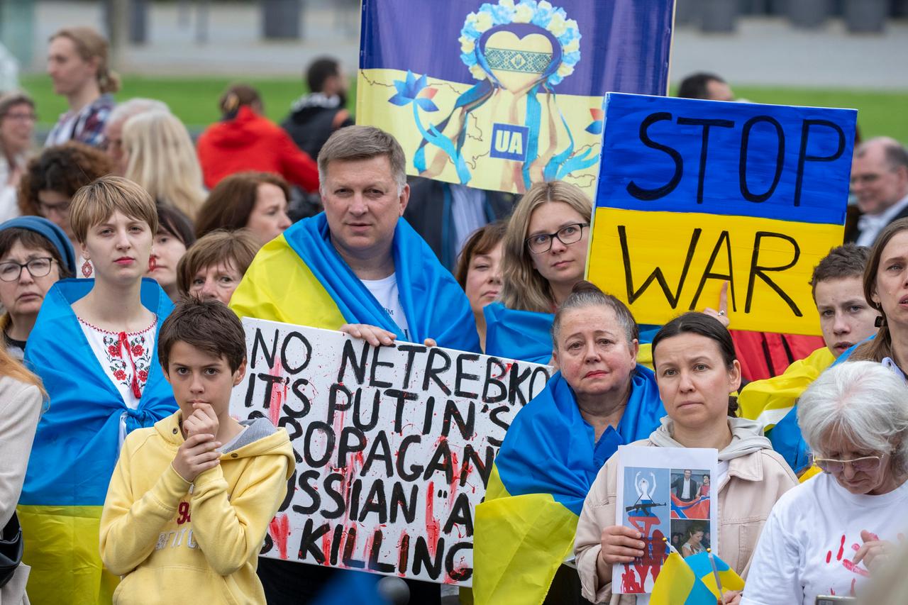 Solidaritätsdemonstration für die Ukraine und gegen einen Auftritt der russischen Sopranistin Anna Netrebko in Wiesbaden. Männer, Frauen und Kinder blicken in Richtung der Kamera. Einige sind in blau-gelbe ukrainische Flaggen gehüllt. Auf einem Schild steht "Stop war", auf Deutsch "Beendet Krieg". Solidaritätsdemonstration für die Ukraine und gegen einen Auftritt der russischen Sopranistin Anna Netrebko in Wiesbaden. Männer, Frauen und Kinder blicken in Richtung der Kamera. Einige sind in blau-gelbe ukrainische Flaggen gehüllt. Auf einem Schild steht "Stop war", auf Deutsch "Beendet Krieg".