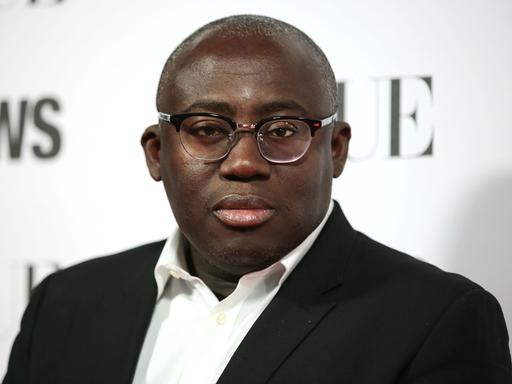 Ein Mann mit dunkler Hautfarbe und Brille steht in schwarzem Jacket und weißem Hemd vor einer weißen Wand mit verschwommenen schwarzen Wörtern