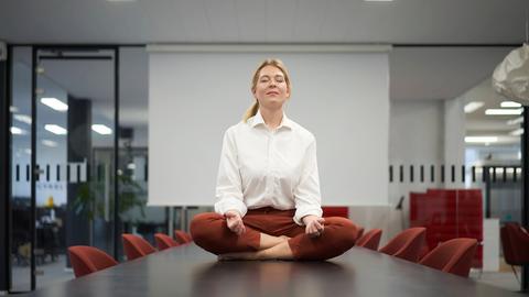 Eine Geschäftsfrau meditiert auf einem Tisch sitzend in einem Konferenzraum. 