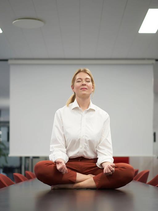 Eine Geschäftsfrau meditiert auf einem Tisch sitzend in einem Konferenzraum. 