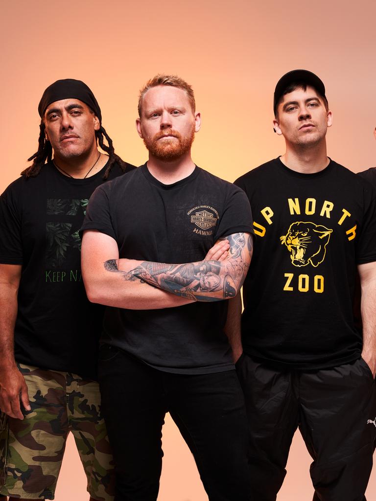 Fünf muskulöse Männer stehen vor einem apricot-farbenen Hintergrund. Sie tragen dunkle Kleidung und blicken ernst. Zusammen sind sie die neuseeländische Reggae-Band L.A.B.