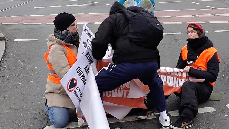 Bei einer Aktion der "Letzten Generation'" blockieren Umweltaktivistinnen eine Kreuzung im Stadtteil Prenzlauer Berg. Ein Mann versucht dabei, ihnen ihre Transparente wegzureißen. Bei einer Aktion der "Letzten Generation'" blockieren Umweltaktivistinnen eine Kreuzung im Stadtteil Prenzlauer Berg. Ein Mann versucht dabei, ihnen ihre Transparente wegzureißen.