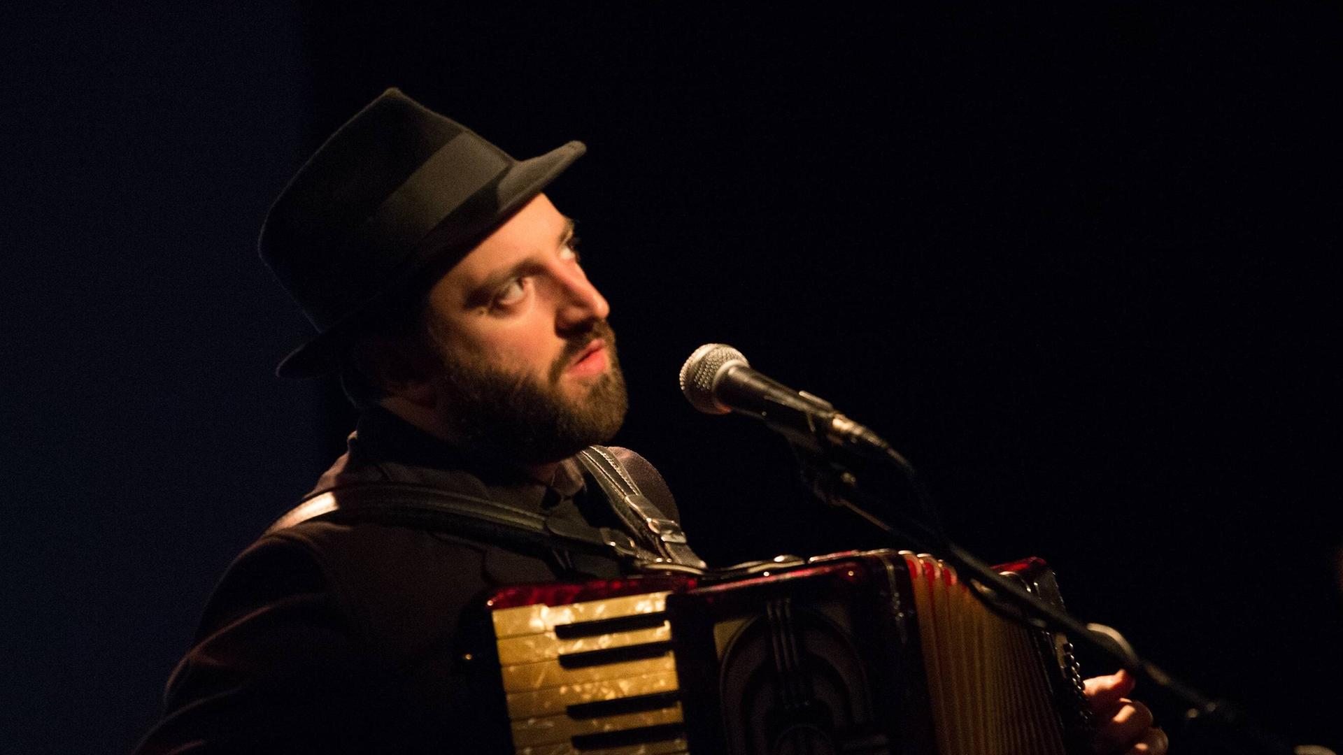 Daniel Kahn mit Hut spielt Akkordeon und singt auf einer Bühne mit Mikrofon. Daniel Kahn mit Hut spielt Akkordeon und singt auf einer Bühne mit Mikrofon.