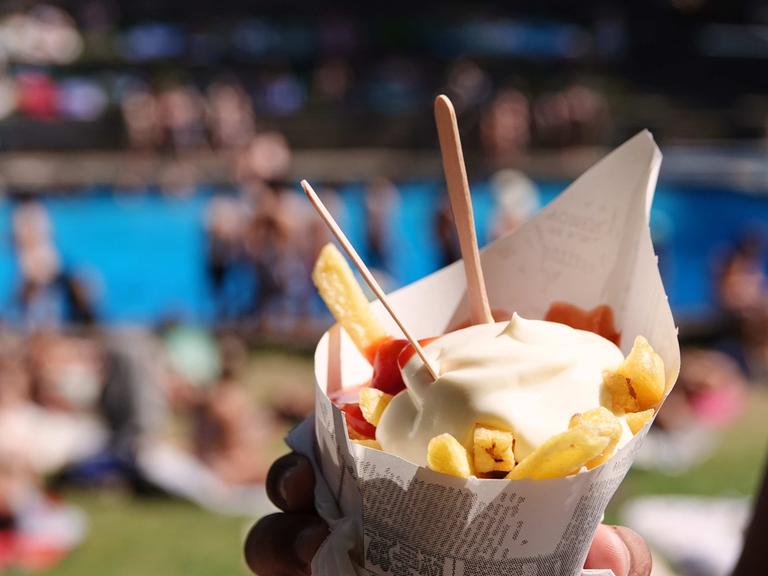 Eine Tüte Pommes Frites, im Hintergrund Menschen in einem Freibad.