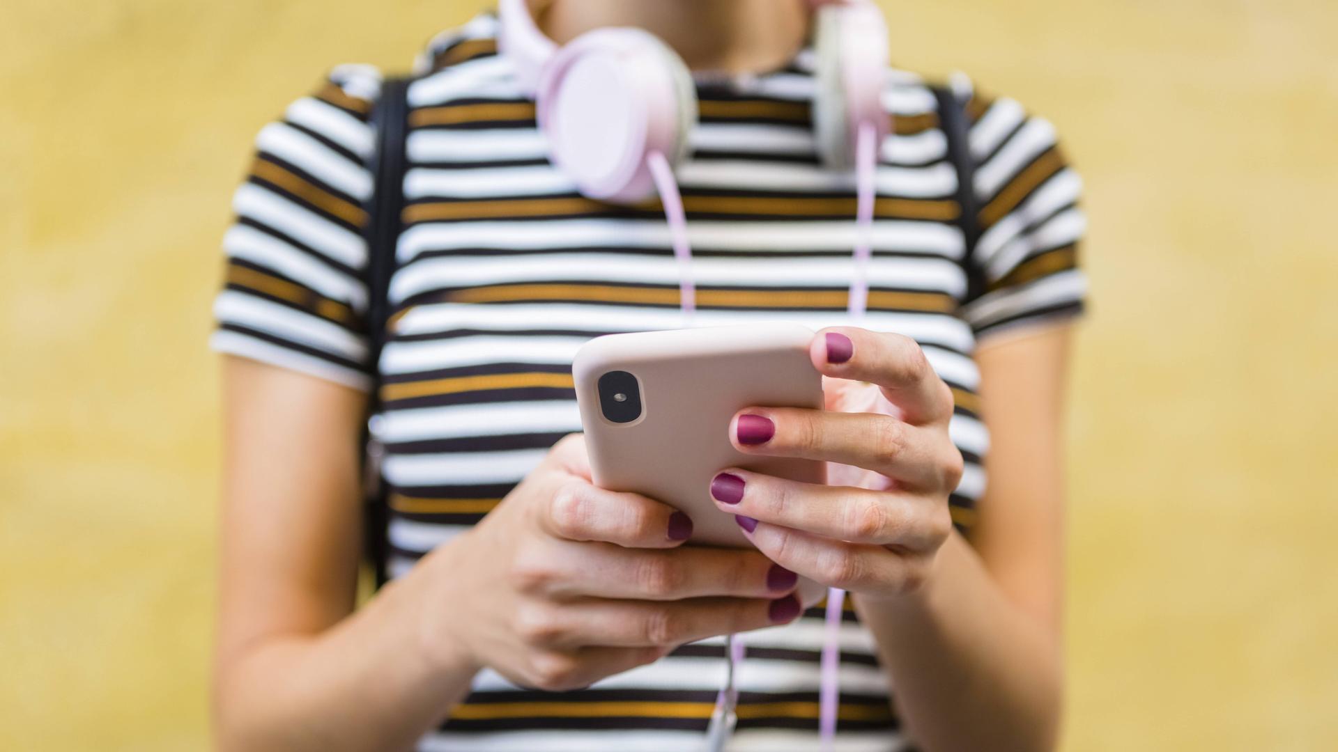 Eine Frau steht mit Smartphone in der Hand und Kopfhörern vor einer gelben Wand Eine Frau steht mit Smartphone in der Hand und Kopfhörern vor einer gelben Wand