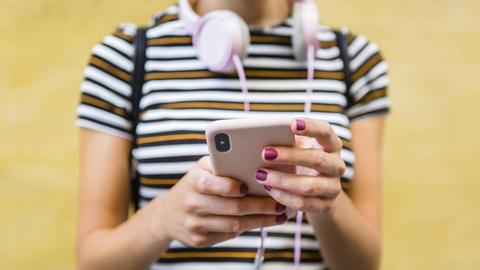Eine Frau steht mit Smartphone in der Hand und Kopfhörern vor einer gelben Wand