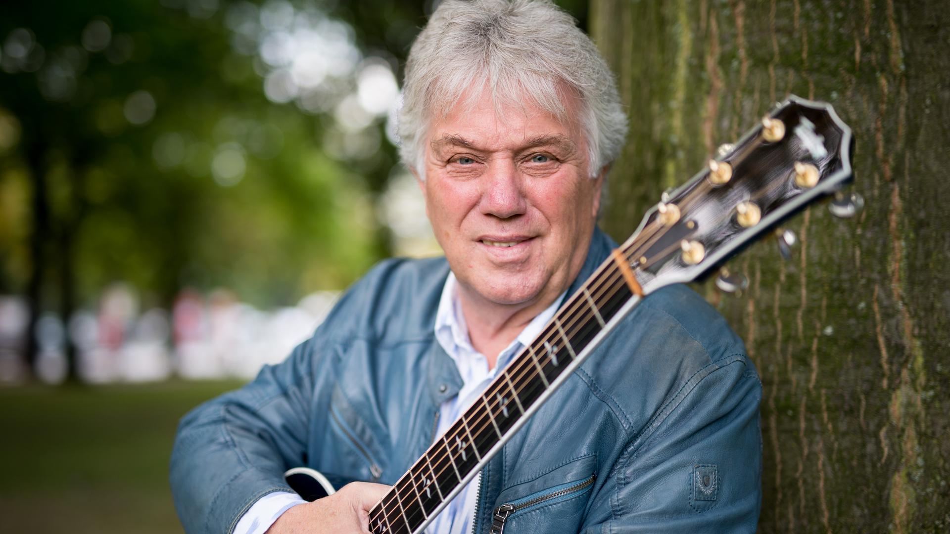 Der Musiker Rolf Zuckowski, mit weißem Haar, in hellgrauer Lederjacke, hält eine Gitarre in der rechten Hand und lehnt an einem Baum.
