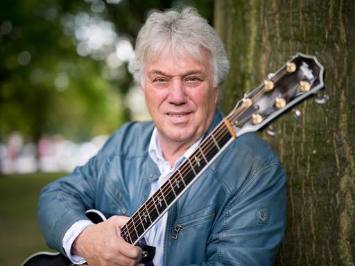 Der Musiker Rolf Zuckowski, mit weißem Haar, in hellgrauer Lederjacke, hält eine Gitarre in der rechten Hand und lehnt an einem Baum.