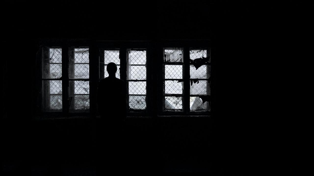 Eine Frau steht als Silhouette im Dunkeln vor vergitterten Fenstern und schaut hinaus Eine Frau steht als Silhouette im Dunkeln vor vergitterten Fenstern und schaut hinaus