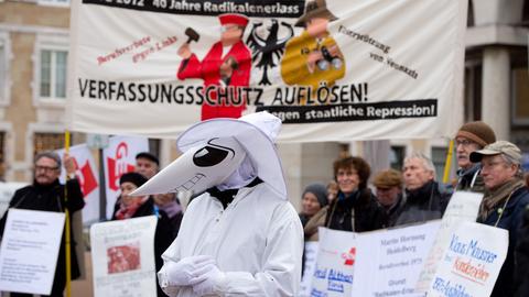 Eine Person mit Maske steht vor einer Menschenmenge mit Protestschildern.