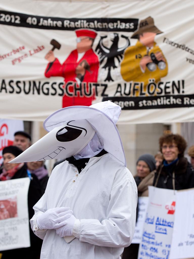 Eine Person mit Maske steht vor einer Menschenmenge mit Protestschildern.