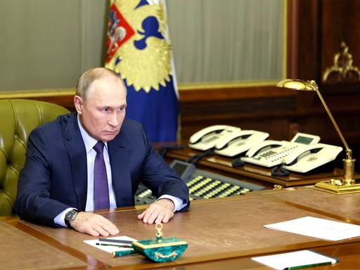 Der russische Präsident Putin, allein an seinem Schreibtisch. Im Hintergrund stehen eine Handvoll Telefone nebeneinander aufgereiht.