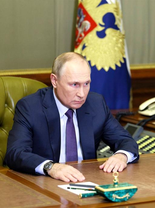 Der russische Präsident Putin, allein an seinem Schreibtisch. Im Hintergrund stehen eine Handvoll Telefone nebeneinander aufgereiht.
