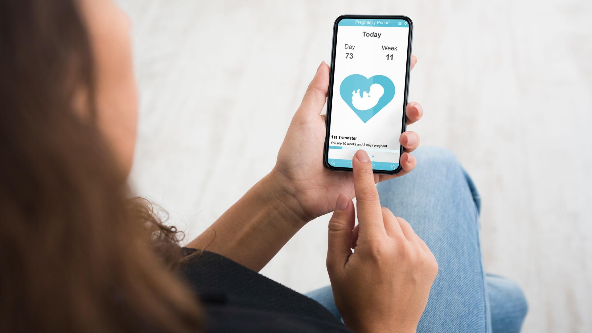 Eine junge Frau in den USA hält ihr Mobiltelefon mit einer Zyklus-App in der Hand, die Illustration von einem Fötus ist auf dem Display zu sehen. Eine junge Frau in den USA hält ihr Mobiltelefon mit einer Zyklus-App in der Hand, die Illustration von einem Fötus ist auf dem Display zu sehen.