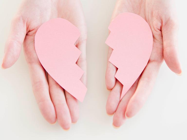 Ein rosafarbenes zerbrochenes Herz aus Kunststoff liegt auf einem Paar Handflächen