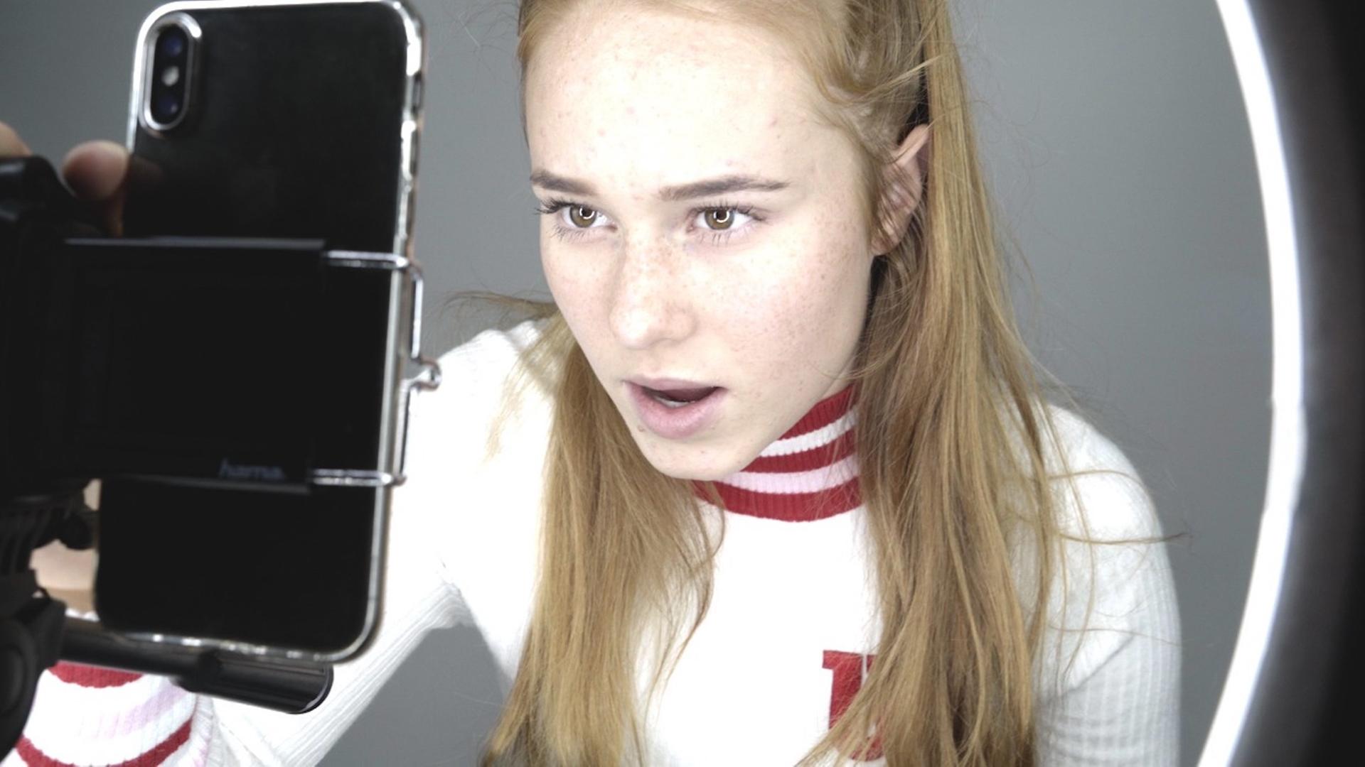 Im Still aus "Girl Gang" spricht ein Mädchen mit langem blonden Haar in die Selfiekamera ihres Smartphones. Im Still aus "Girl Gang" spricht ein Mädchen mit langem blonden Haar in die Selfiekamera ihres Smartphones.