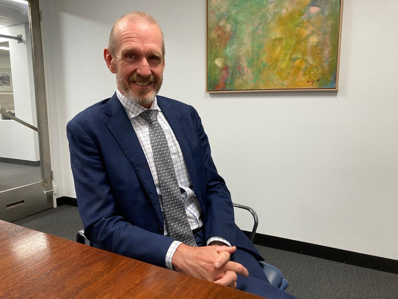 Ein älterer Herr mit lichtem Haar, blauem Jacket und grauer Krawatte sitzt am Tisch im Büro vor einem großen bunten Bild. Ein älterer Herr mit lichtem Haar, blauem Jacket und grauer Krawatte sitzt am Tisch im Büro vor einem großen bunten Bild.