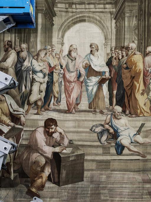 Detail der Anbringung eines Wanteppichs. Links im Bild ist eine Hebebühne zu sehen, zwei Arbeiter stützen den Wanteppich mit Stangen.
Das Motiv zeigt einen monumentalen Innenraum, in dem eine Versammlung maßgeblicher Wissenschaftler und Philosophen von der Antike bis zur Renaissance stattfindet. 