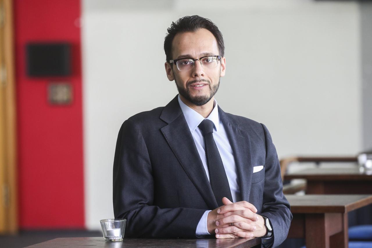 Muhammad Sameer Murtaza, mit Brille, dunklem Sakko und schwarzer Krawatte, steht an einem Tisch und hat die Hände gefaltet. Muhammad Sameer Murtaza, mit Brille, dunklem Sakko und schwarzer Krawatte, steht an einem Tisch und hat die Hände gefaltet.