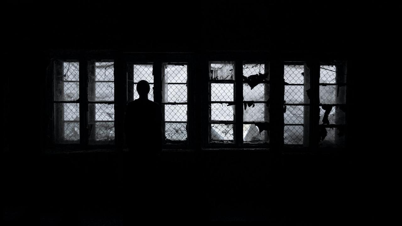 Eine Frau steht als Silhouette im Dunkeln vor vergitterten Fenstern und schaut hinaus Eine Frau steht als Silhouette im Dunkeln vor vergitterten Fenstern und schaut hinaus