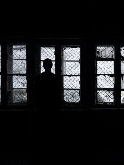 Eine Frau steht als Silhouette im Dunkeln vor vergitterten Fenstern und schaut hinaus