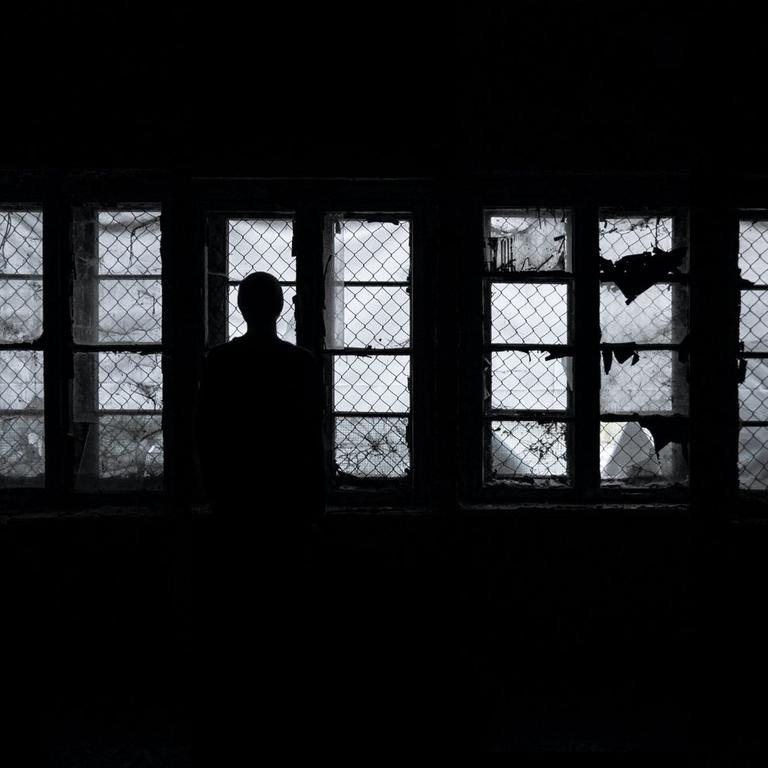 Eine Frau steht als Silhouette im Dunkeln vor vergitterten Fenstern und schaut hinaus Eine Frau steht als Silhouette im Dunkeln vor vergitterten Fenstern und schaut hinaus
