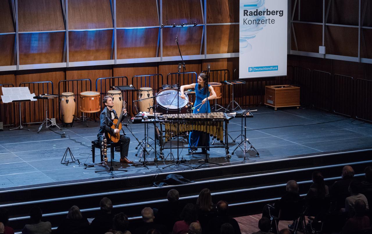 Ein Gitarrist und eine Perkussionistin musizieren auf einer Bühne vor Publikum. Ein Gitarrist und eine Perkussionistin musizieren auf einer Bühne vor Publikum.
