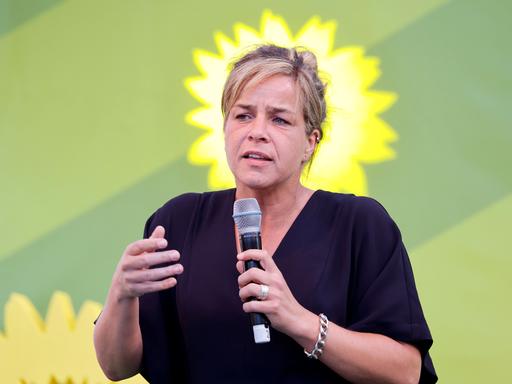 Ein Frau spricht in ein Mikrofon, im Hintergrund sind die Umrisse der gelben Sonnenblume der Grünen zu sehen.