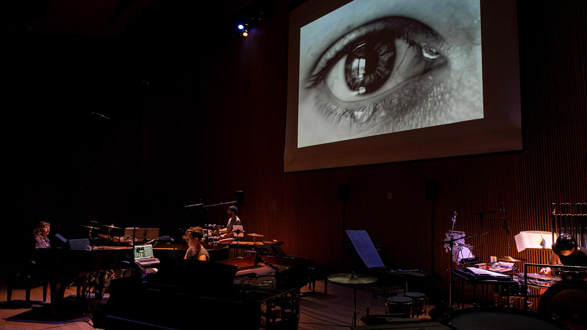 Das Ensemble Yarn/Wire spielt auf der Bühne während in der Videoprojektion oberhalb ein tränens Auge zu sehen ist Das Ensemble Yarn/Wire spielt auf der Bühne während in der Videoprojektion oberhalb ein tränens Auge zu sehen ist