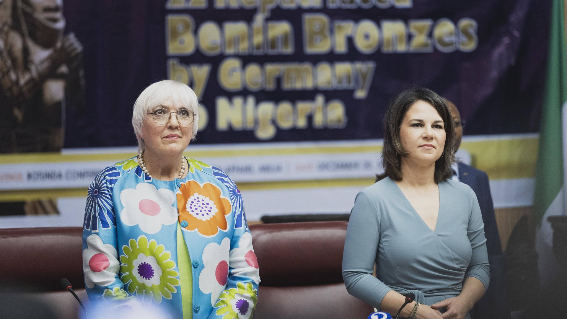Claudia Roth und Annalena Baerbock stehen bei der feierlichen Übergabe von Benin-Bronzen an den nigerianischen Staat im Dezember 2022 nebeneinander und hören eine Rede zu.