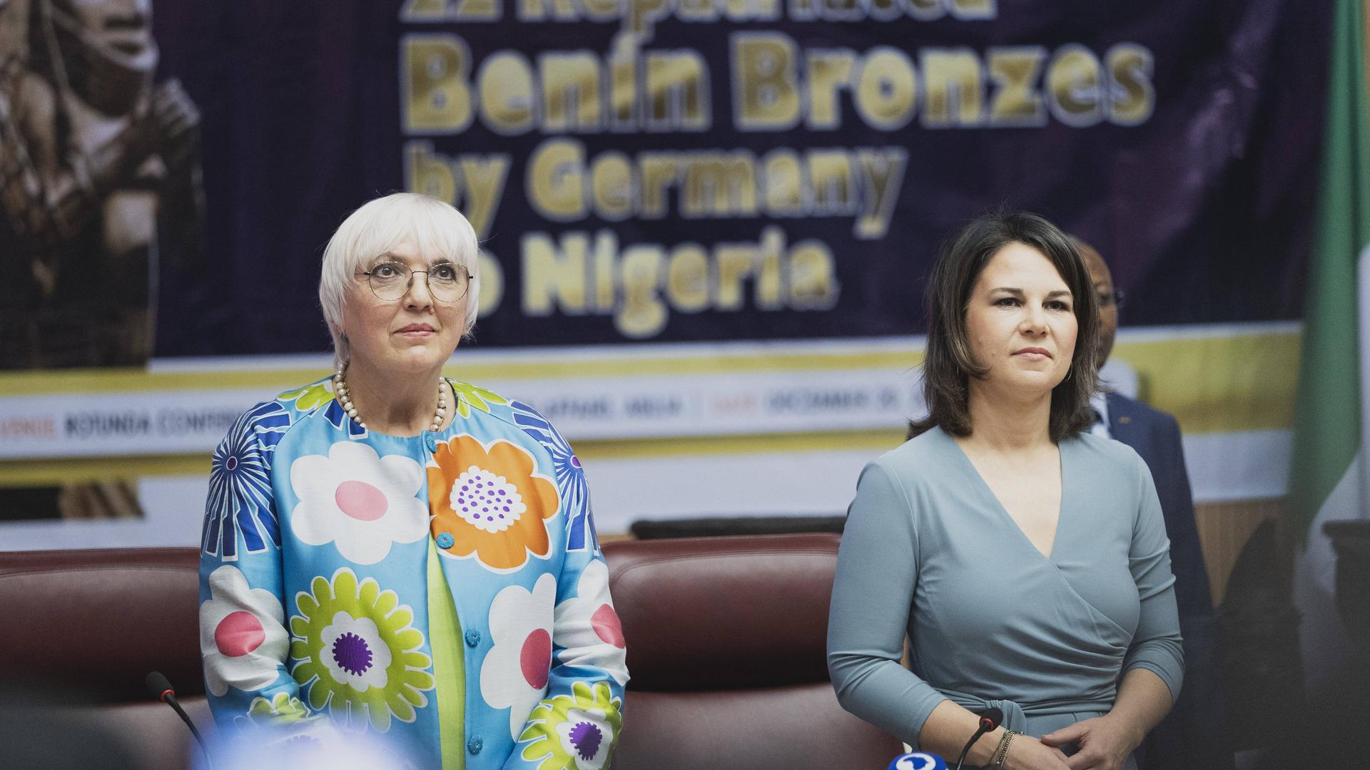Claudia Roth und Annalena Baerbock stehen bei der feierlichen Übergabe von Benin-Bronzen an den nigerianischen Staat im Dezember 2022 nebeneinander und hören eine Rede zu. Claudia Roth und Annalena Baerbock stehen bei der feierlichen Übergabe von Benin-Bronzen an den nigerianischen Staat im Dezember 2022 nebeneinander und hören eine Rede zu.