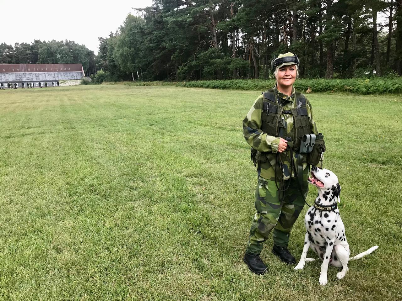 Eine junge Frau in militärischer Tarnkleidung steht mit einem Dalmatiner-Hund auf einer Wiese.