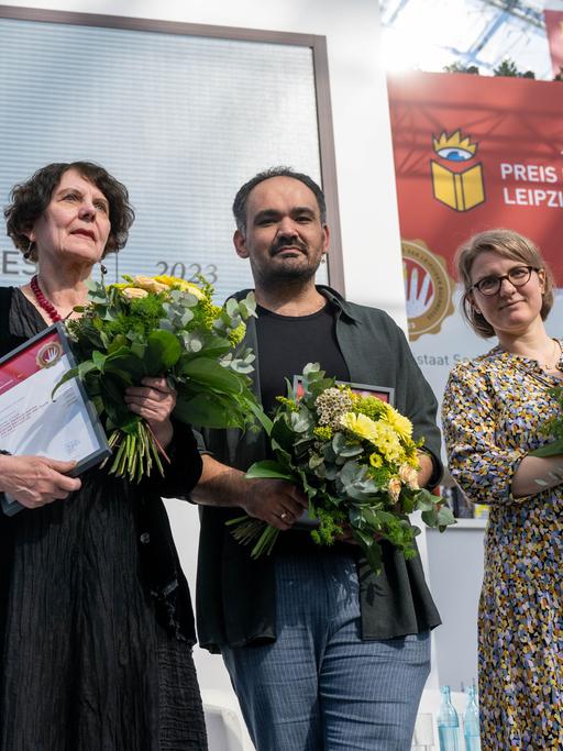 Regina Scheer, Dinçer Güçyeter und Johanna Schwering (v.l.) stehen mit Blumensträußen auf der Bühne.