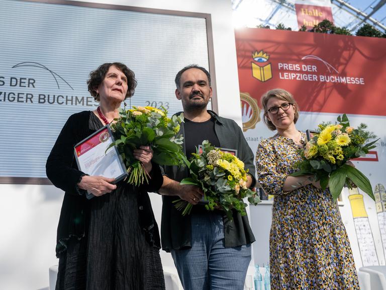 Regina Scheer, Dinçer Güçyeter und Johanna Schwering (v.l.) stehen mit Blumensträußen auf der Bühne.