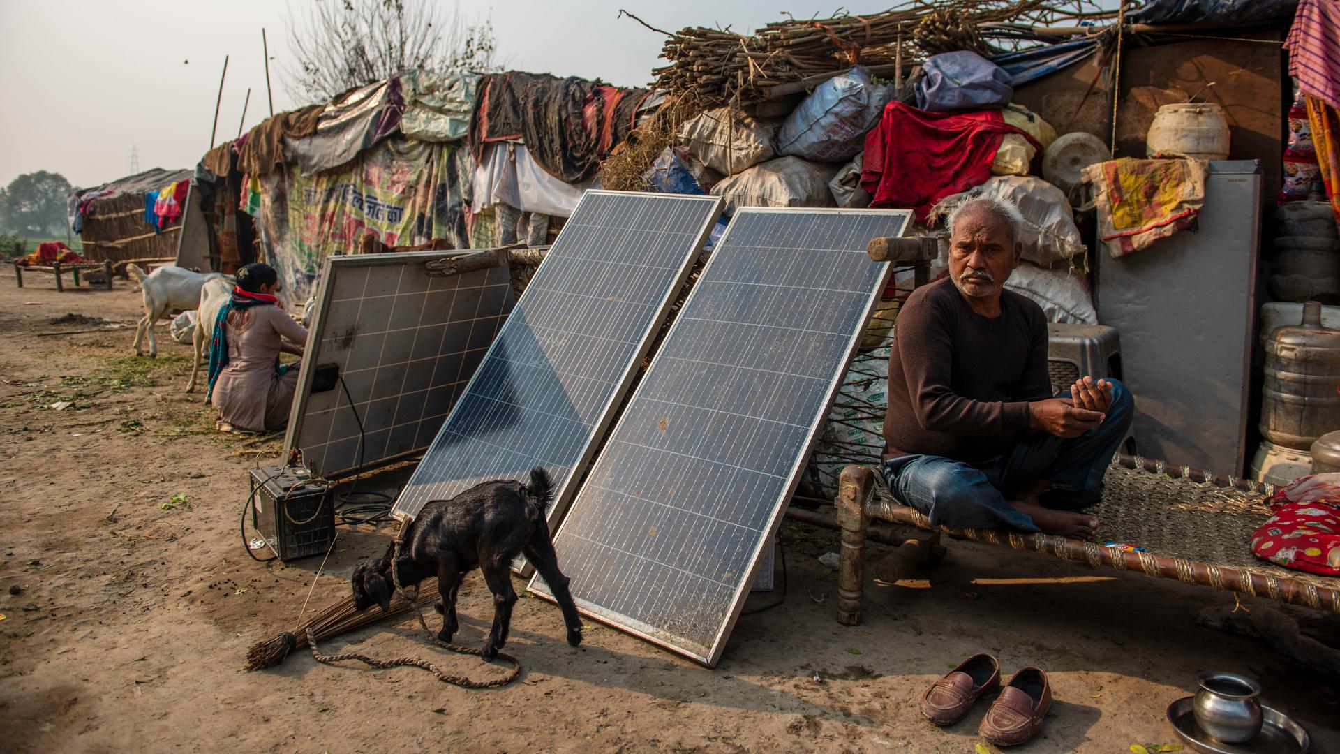 Ein Mann sitzt vor einer zeltartigen, einfachen Hütte, neben ihm drei Solar-Panels und ein kleiner Generator, vor denen eine kleine Ziege angebunden ist, die auf dem staubigen Boden schnuppert. Ein Mann sitzt vor einer zeltartigen, einfachen Hütte, neben ihm drei Solar-Panels und ein kleiner Generator, vor denen eine kleine Ziege angebunden ist, die auf dem staubigen Boden schnuppert.