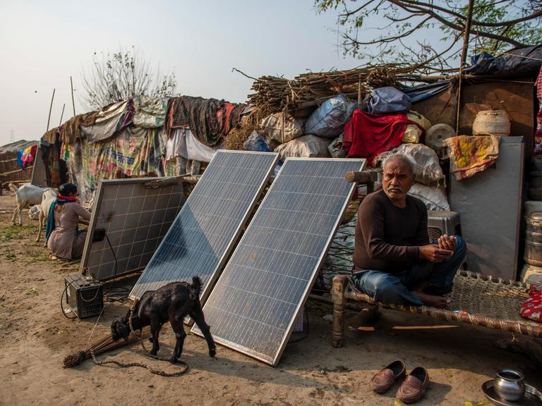 Ein Mann sitzt vor einer zeltartigen, einfachen Hütte, neben ihm drei Solar-Panels und ein kleiner Generator, vor denen eine kleine Ziege angebunden ist, die auf dem staubigen Boden schnuppert.