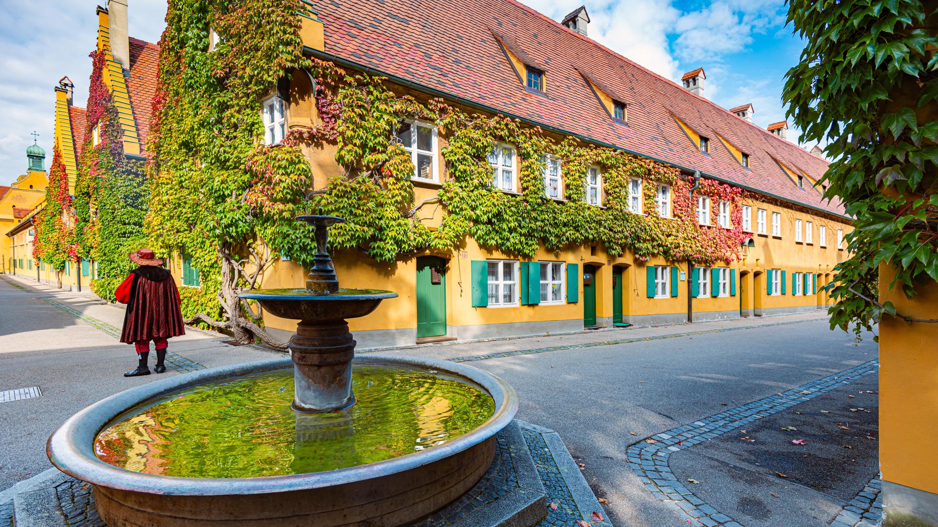 Eine sommerliche Aufnahme zeigt die „Fuggerei“ in Augsburg: zweigeschossige, orange gestrichene Häuser.