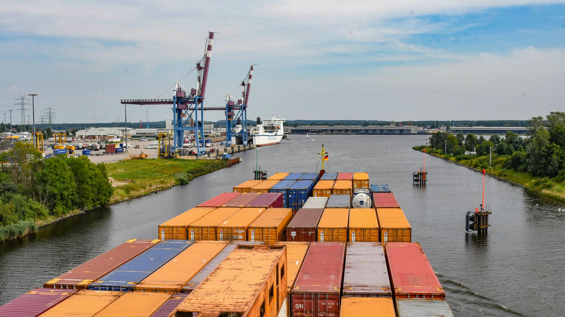 Im Vordergrund sind Container auf einem Frachtschiff zu sehen, im Hintergrund Kräme am Ufer sowie die Trave, auf der das Schiff fährt. Im Vordergrund sind Container auf einem Frachtschiff zu sehen, im Hintergrund Kräme am Ufer sowie die Trave, auf der das Schiff fährt.