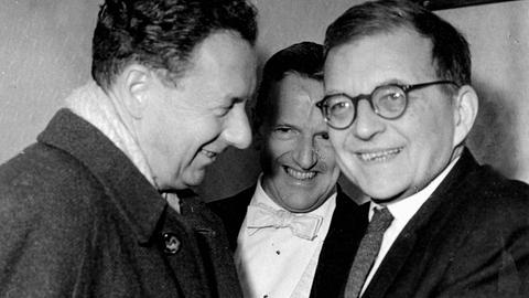 Benjamin Britten und Dmitri Schostakowitsch schütteln einander die Hände, während ein dritter Mann zwischen ihnen die Szene lächelnd beobachtet. 
